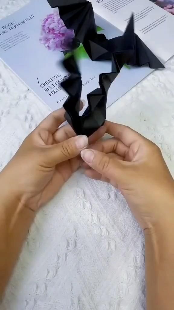 two hands holding black origami pieces on top of a white sheet with pink flowers in the background