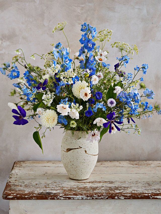 a white vase filled with blue and white flowers on top of a wooden table next to a wall