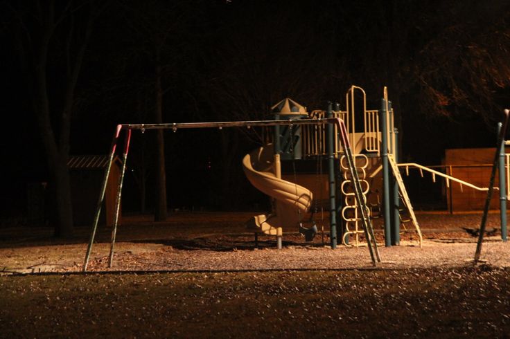 an empty playground at night with a slide and climbing frame in the foreground, lit by street lights