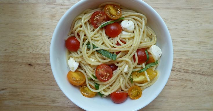 a white bowl filled with pasta, tomatoes and mozzarella on top of a wooden table