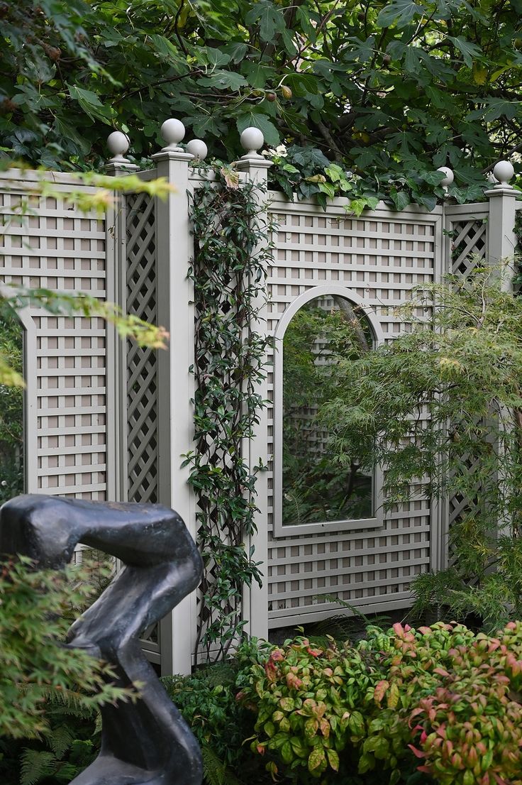 a statue in the middle of a garden with trees and bushes around it, next to a white trellis