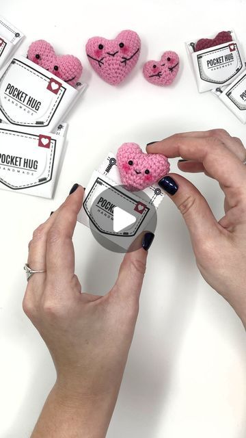 two hands are holding small pink crocheted teddy bears in front of some packaging