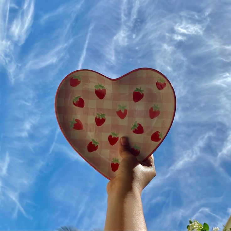 someone holding up a heart shaped plate with strawberries on it in front of a blue sky