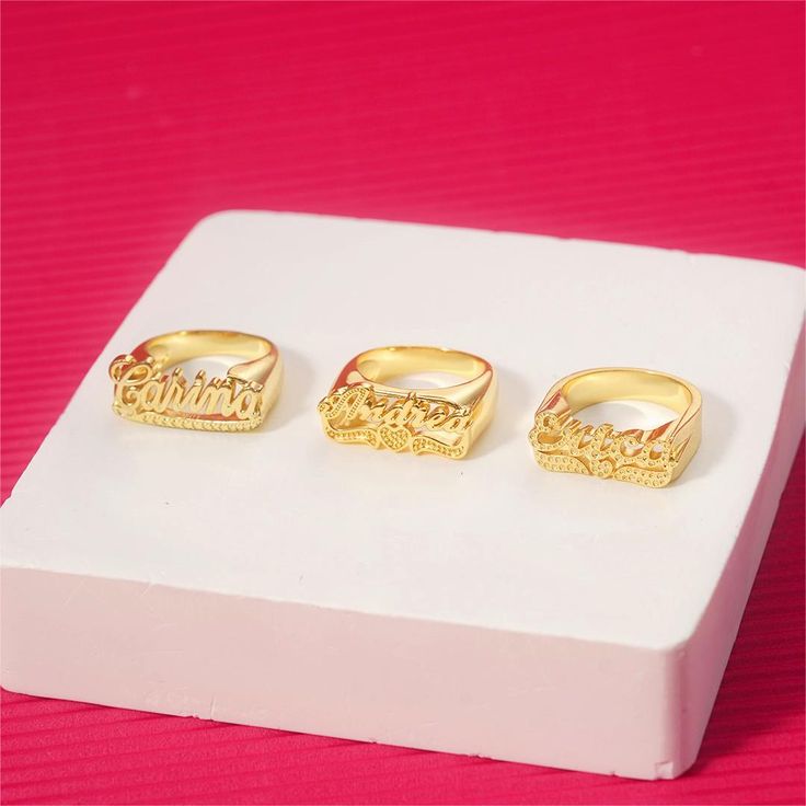 three gold rings sitting on top of a white display box with pink background and red tablecloth