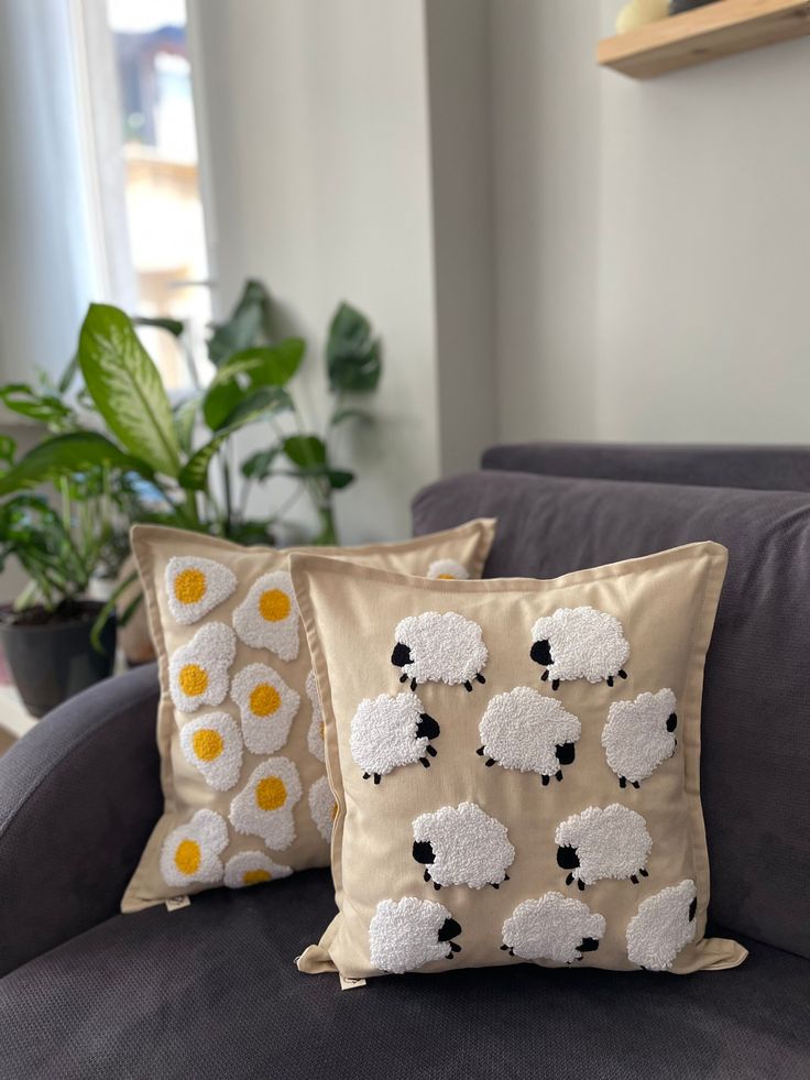 two pillows with white sheep on them sitting on a couch in front of a potted plant