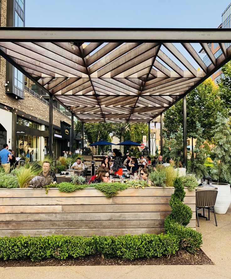 people are sitting on benches under an awning in a city park area with trees and bushes