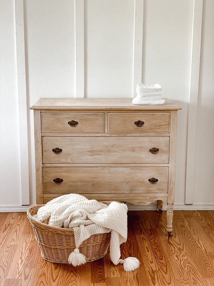 a basket with blankets sitting on top of it next to a wooden dresser and white wall