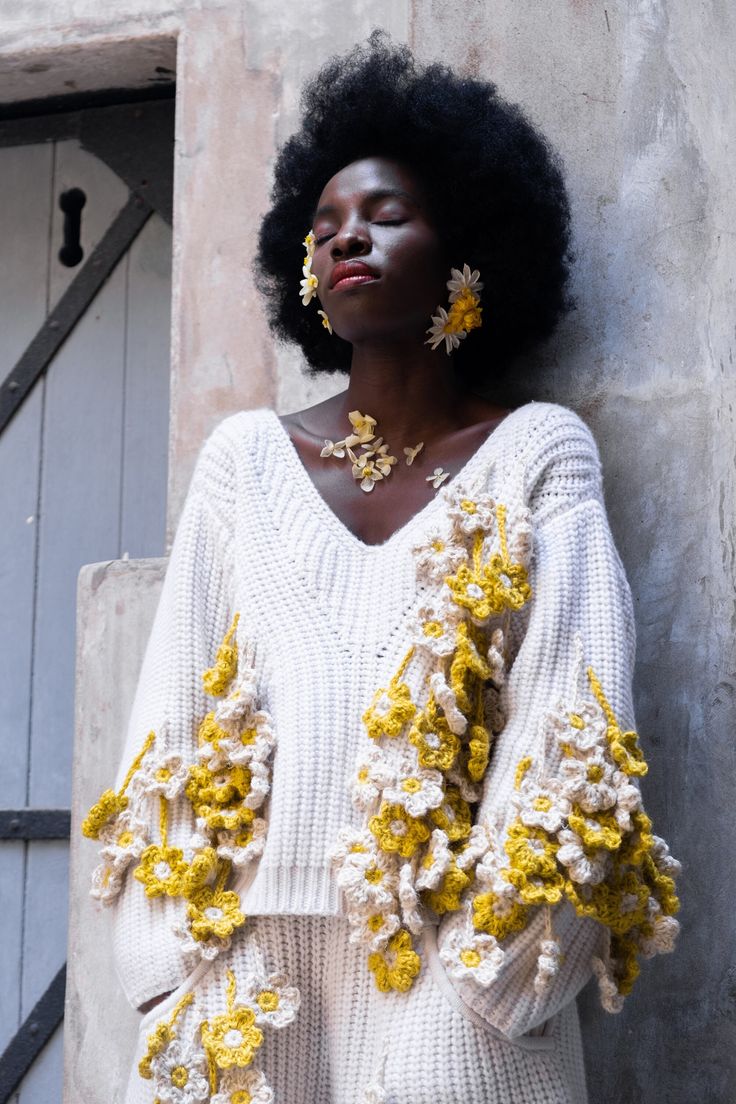 a woman standing next to a building with flowers on her head and sweater over her shoulders