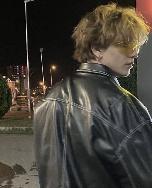 a young man wearing sunglasses and a black leather jacket standing next to a street sign