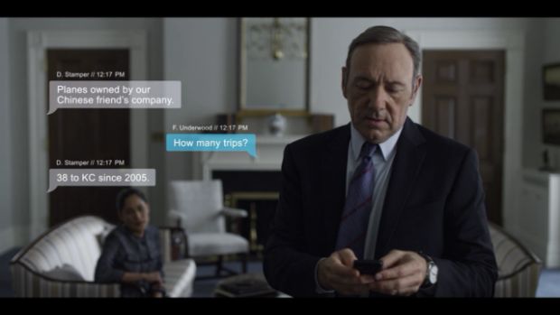 a man in a suit and tie looking at his cell phone with another person sitting on the couch behind him