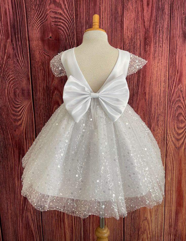 Sequin Pearl Dress This beautiful elegant dress is handmade, bodice is made of bridal satin with a short beaded cap sleeve, the back of the dress consists of a deep V-back with a zipper. The big bow has a pin to be attached to the dress and it's removable if needed. The skirt consists of multi layers, the top layer consists of sequin & pearls, 3 layers of tulle, 2 layers of lining with crinoline for fullness.  Dress Is Pictured with a petticoat NOT INCLUDED https://www.etsy.com/listing/1316417982/white-knee-length-petticoat-wedding?click_key=41cb8b0ae4587efa582b0a433f3f5e0cde12aec0%3A1316417982&click_sum=af1df37b&ref=shop_home_active_1 Matching hair bow https://www.etsy.com/listing/1405032366/satin-big-bow-hair-clip-flower-girl?click_key=91c1fea2f7cd1e7b097d731e925a67b789621873%3A140503236 White Princess Dress With Satin Bow For Baptism, Fitted Princess Dress With Satin Bow For First Communion, Tulle Baptism Dress With Satin Bow For First Communion, White Tulle Baptism Dress With Satin Bow, White Baptism Dress With Satin Bow, Fitted Princess Dress With Satin Bow For Baptism, White Satin Princess Dress For Baptism, Fitted White Organza Pageant Dress, White First Communion Dress With Satin Bow For Party