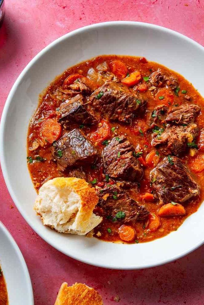 a white bowl filled with meat and carrots next to bread on a pink surface