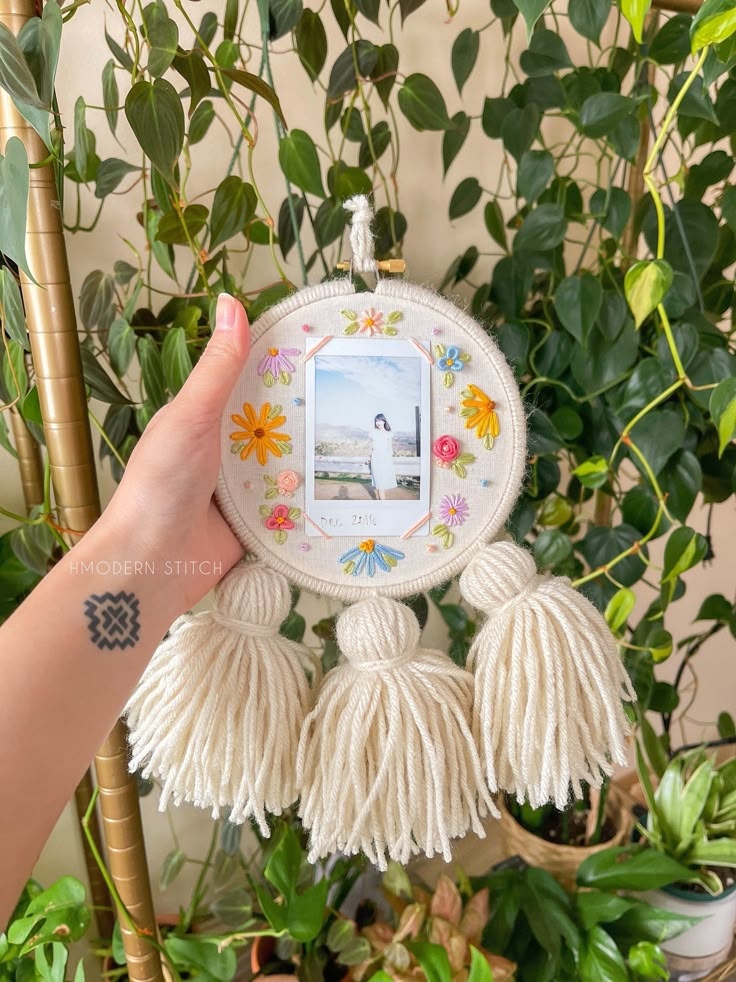 a person holding up a small photo frame with tassels on it in front of some plants