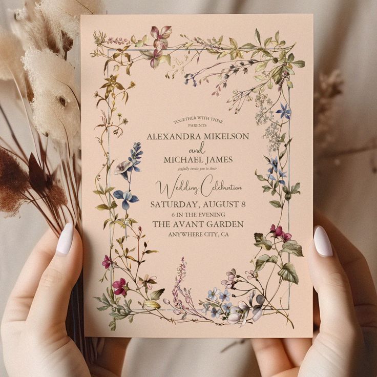 two hands holding up a wedding card with floral designs on the front and back, along with dried flowers