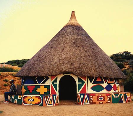 a thatched hut with colorful designs painted on the side and a woman standing in front