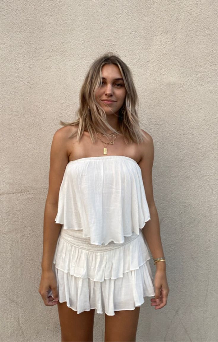 The Monaco White Strapless Romper is a vacay dream! White woven rayon fabric falls from a strapless neckline, with no-slip strips, to a flowy, ruffle overlay bodice with an on-trend smocked overlap back detail. Attached shorts complete the look. Bandeau Tube Top With Smocked Back For Beach, Chic Bandeau Smocked Top For Vacation, Vacation Bandeau Smocked Top, Chic Strapless Smocked Top For Beach, Summer Strapless Smocked Top With Ruffles, Chic Smocked Top With Elastic Neckline For Vacation, Summer Bandeau Tube Top With Smocked Back, Chic Bandeau Tube Top For Brunch, Beach Bandeau Smocked Top With Ruffles