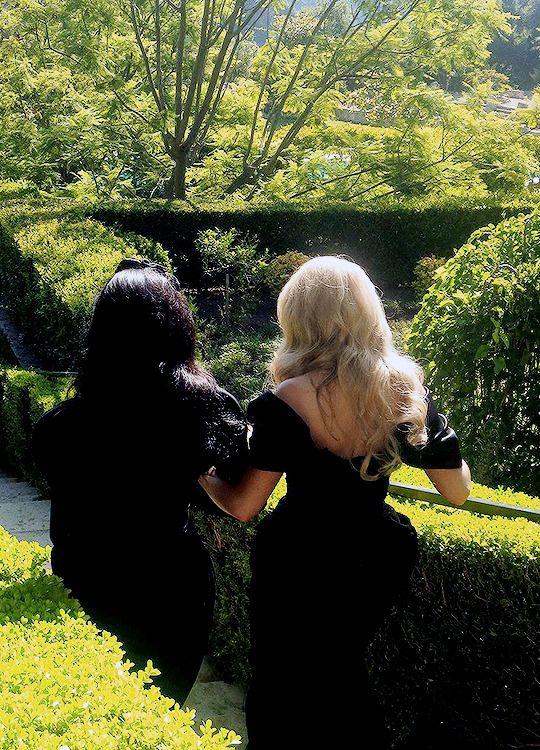 two women in black dresses are walking through the bushes and trees, one is looking at another woman with her back to the camera