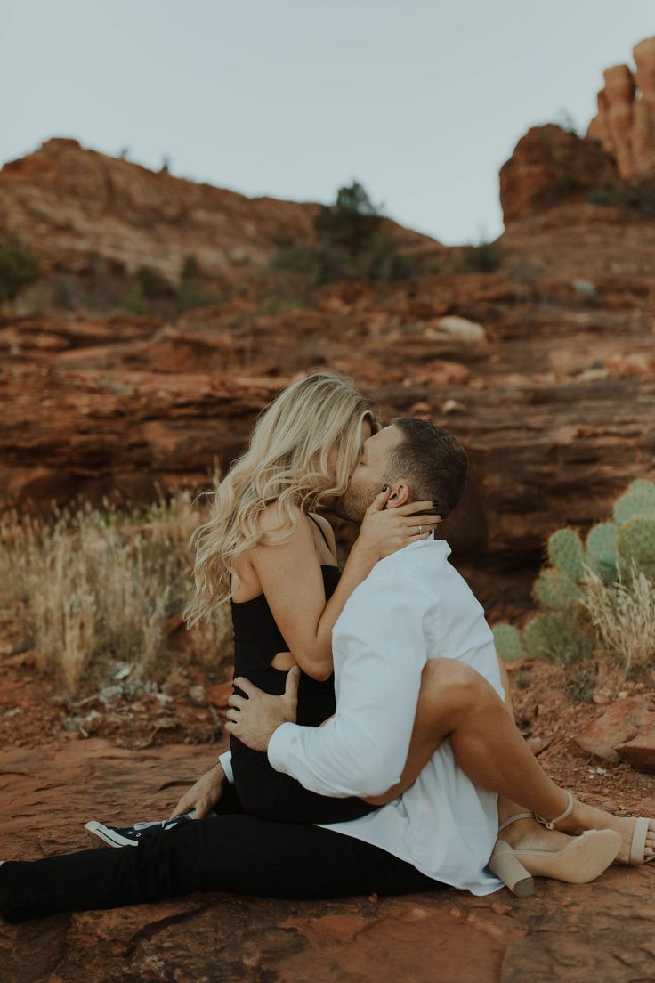 a man and woman sitting on the ground in front of some rocks with their arms around each other