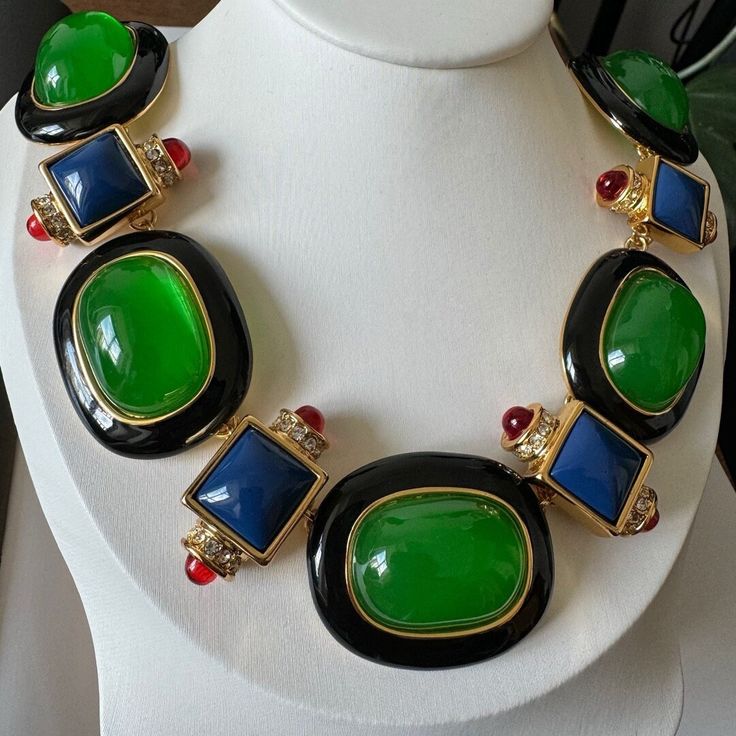 Elevate your style with this luxurious statement necklace. Featuring an exquisite combination of bold green and deep blue stones, this piece is designed to captivate attention. The large oval green stones, set in polished gold-tone metal and framed by glossy black enamel, serve as the striking centerpiece. The square-cut blue stones, adorned with sparkling clear rhinestones and accented by smaller red cabochons, add a touch of geometric elegance to the design. Crafted with meticulous attention t Luxury Statement Jewelry With Oval Cabochon, Luxury Multicolor Artistic Necklaces, Luxury Multicolor Necklace With Large Pendant, Luxury Green Oval Necklace, Formal Green Stone Necklaces, Formal Green Stone Necklace, Luxury Green Stone Necklaces, Luxury Green Emerald Necklace With Stones, Green Oval Jewelry For Party