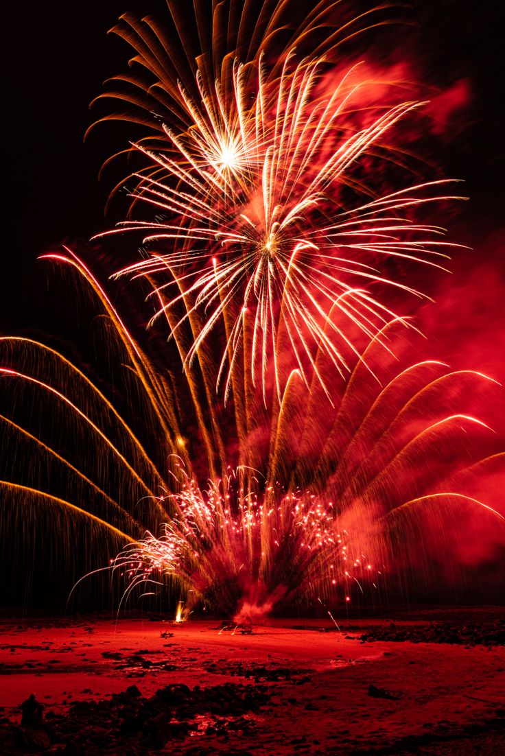 fireworks are lit up in the night sky over water and beach with red, yellow, and green colors