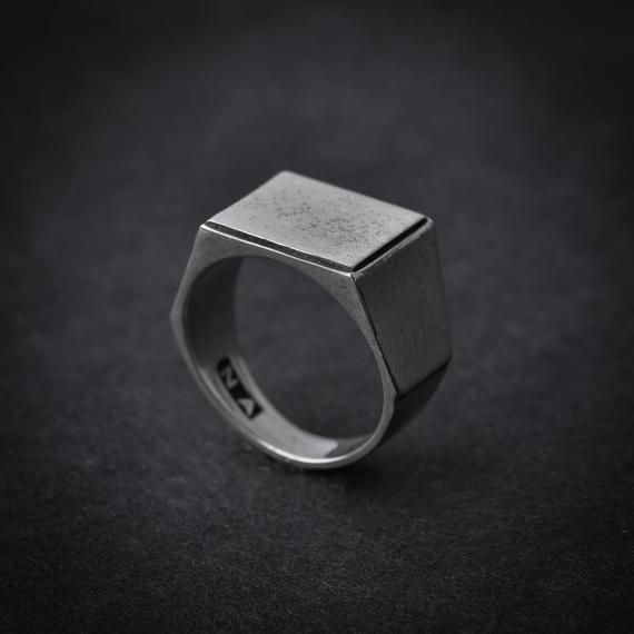 a silver ring with a square shaped design on the front and center, sitting on a black surface