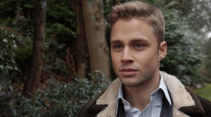 a man in a jacket and tie looking at the camera while standing next to some trees