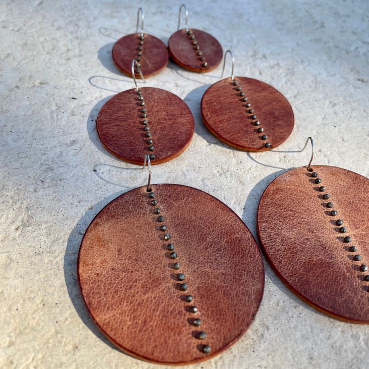 Distressed chestnut brown leather disks, with hand-sewn pyrite beads, hang from surgical steel ear wires. Three sizes and so light, you'll forget you're wearing them. Small; 1.75" L x 1.25" W Medium: 2.5L x 1.75W Large: 3"L x 2.25"W Leather And Metal Earrings, Simple Leather Earrings, Handmade Leather Earrings Ideas, Handmade Circular Brown Jewelry, Handmade Brown Circular Jewelry, Handmade Rust Leather Jewelry, Brown Leather Jewelry With Soldered Details, Unique Hand Forged Brown Earrings, Unique Brown Round Earrings