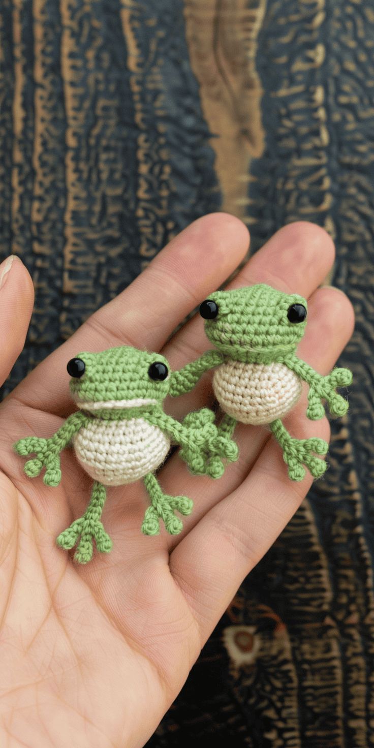 two small crocheted frog figurines sitting on top of each other in the palm of someone's hand