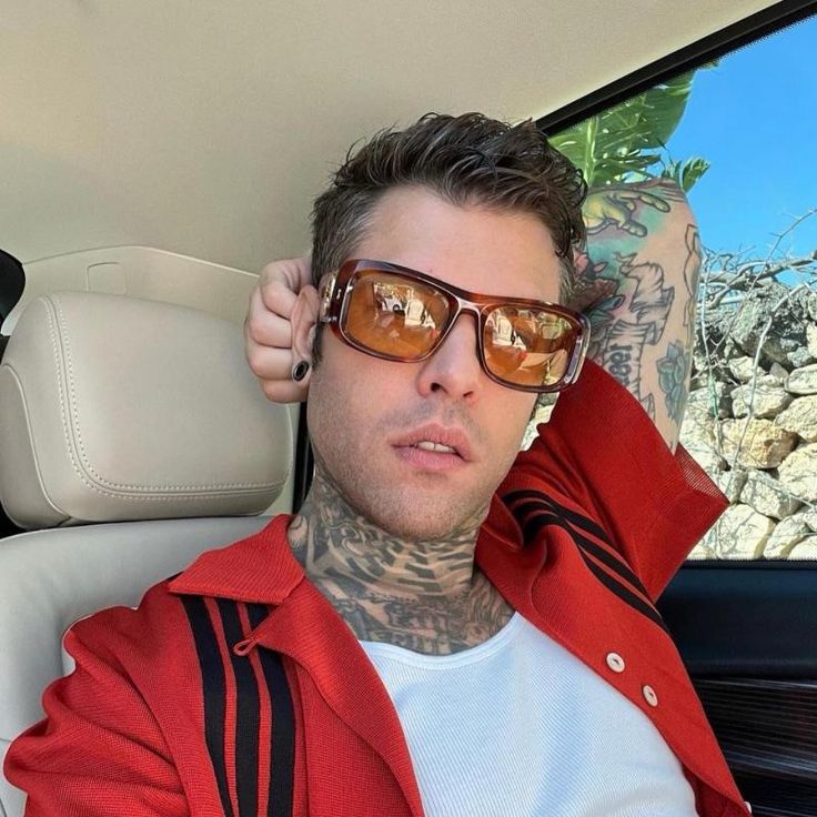 a man with tattoos sitting in the back seat of a car wearing sunglasses and a red jacket