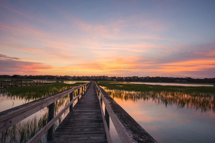 a wooden bridge over water at sunset with grass in the foreground and clouds in the background