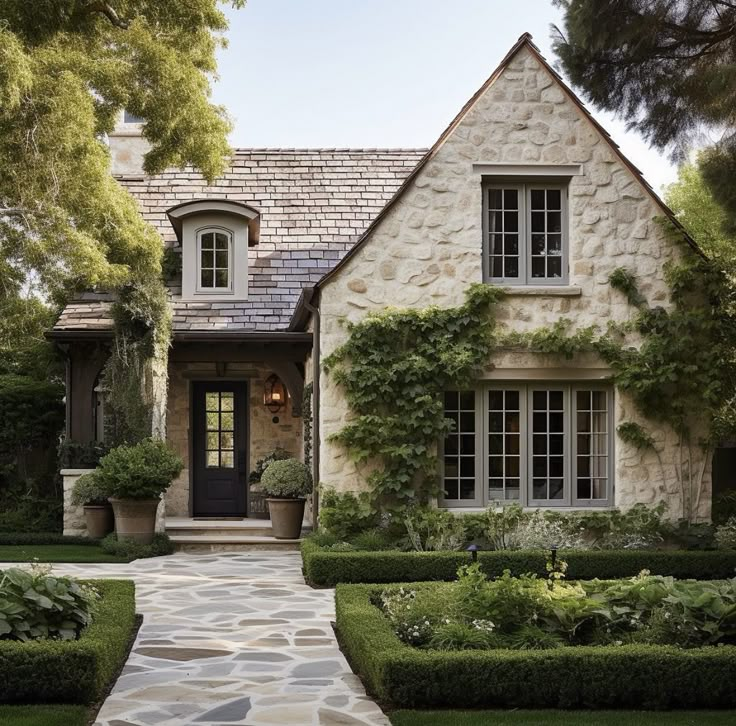 a stone house with trimmed hedges and potted plants in the front yard, surrounded by greenery