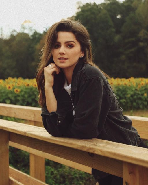 a woman is sitting on a wooden bench in front of sunflowers and trees