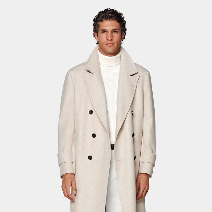 Mid-thigh length overcoat in sand, featuring wide peak lapels and natural shoulders. Double-breasted closure, patch pockets, and inside pockets for functionality. Single vent, lined, with martingale belt for style. Luxury Cream Outerwear With Double Button Closure, Beige Wool Shawl Collar Outerwear, Luxury Beige Gabardine Outerwear, Neutral Long Wool Coat, Double-breasted Cream Wool Outerwear, Cream Wool Coat For Business In Winter, Cream Outerwear With Concealed Placket For Fall, Beige Notch Lapel Wool Coat For Winter, Cream Double-breasted Wool Outerwear