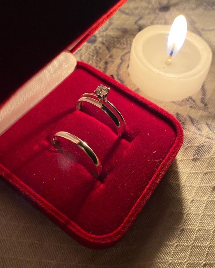 two wedding rings in a red velvet box next to a candle