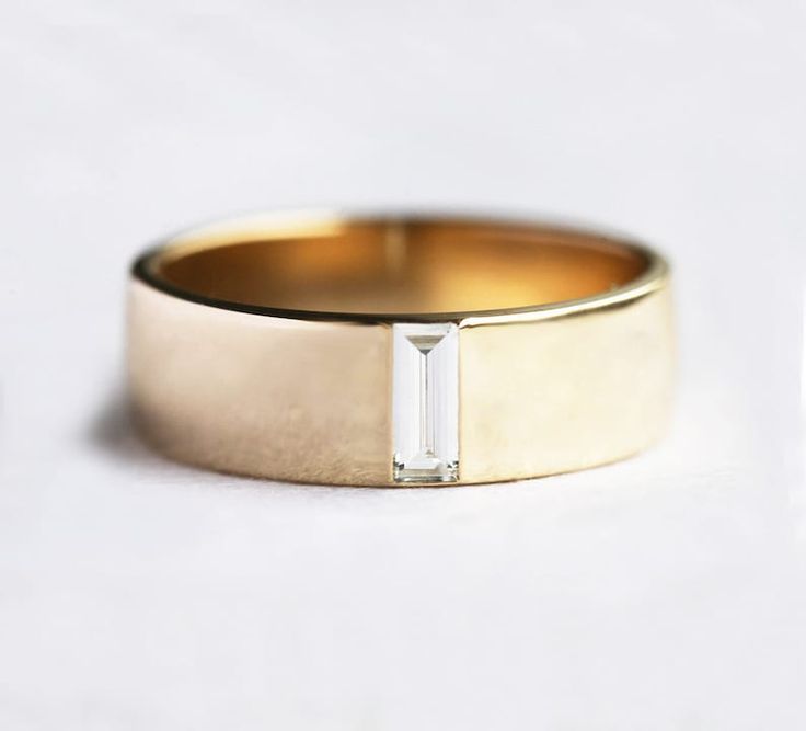 a gold wedding band with a baguette cut diamond in the center, on a white surface