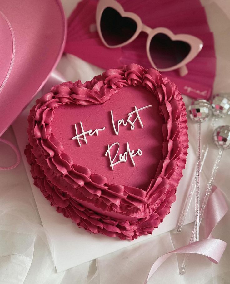 a heart shaped cake with the words her last love written on it next to pink hats and sunglasses