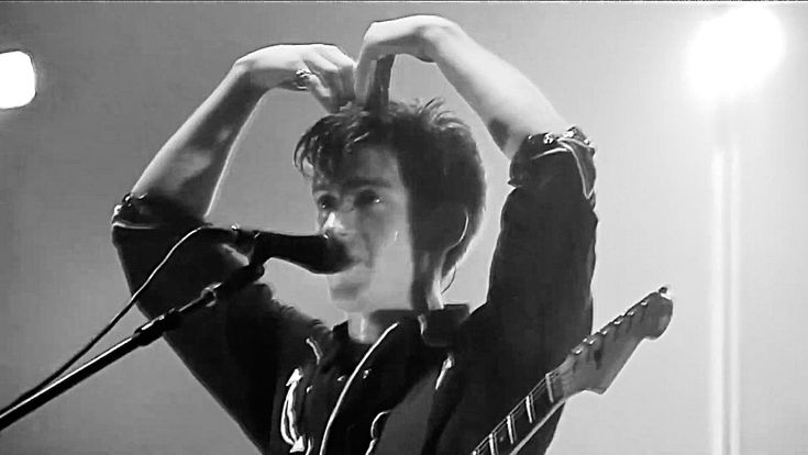 a young man holding his guitar up to his head while singing into a microphone in front of him