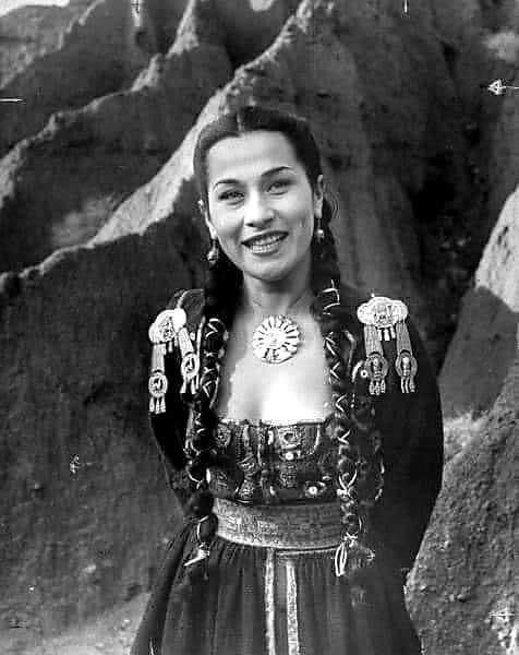 an old black and white photo of a woman in native dress standing next to some rocks