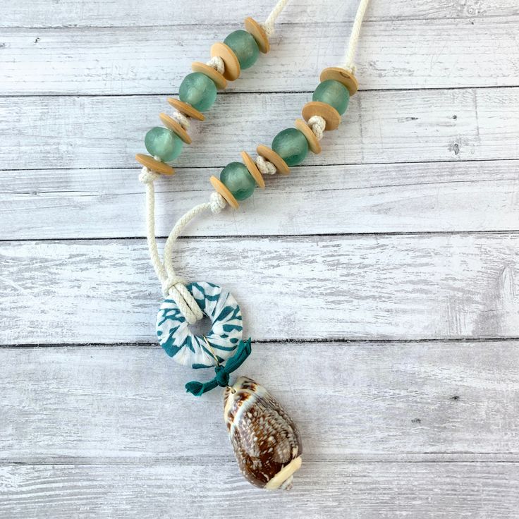 Beach-themed necklace that is ready to jump in your suitcase for that beach vacation! A 2-Inch seashell from an Indian Ocean Mix of shells hangs from a fabric-wrapped wooden hoop with a little bow made of vintage sari silk. This seashell and hoop pendant measuring 4-Inches sits center stage on this beach-inspired pendant necklace complimented with sage sea glass beads and wooden saucer spacer beads strung on 1/8-inch wide rope cord with brass end caps and lobster claw closure. This calming palet Ocean-inspired Green Necklaces For Beach, Green Ocean-inspired Necklaces For The Beach, Green Ocean-inspired Necklaces For Beach, Unique Beaded Necklaces With Wooden Beads For Beach, Green Ocean-inspired Beach Necklaces, Handmade Ocean Color Necklaces For Beach, Handmade Coastal Beach Necklaces, Handmade Coastal Style Necklaces For Beach, Handmade Coastal Necklaces For Vacation