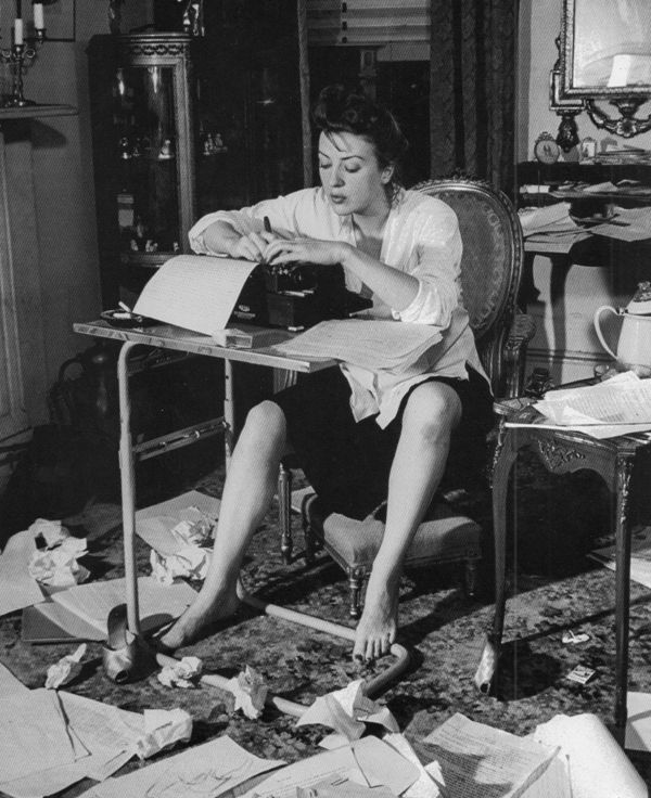 a woman sitting at a desk with papers all over the floor and in front of her