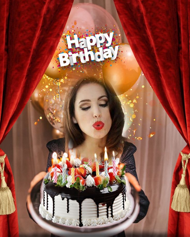 a woman holding a birthday cake with lit candles on it in front of red curtains