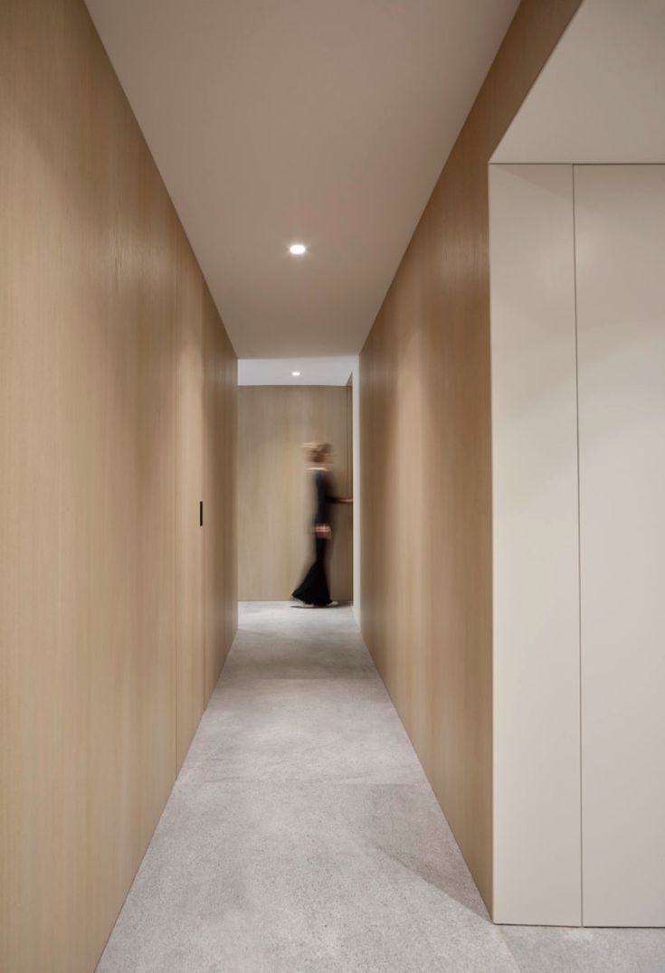 a woman walking down a long hallway with white carpet on the floor and beige walls