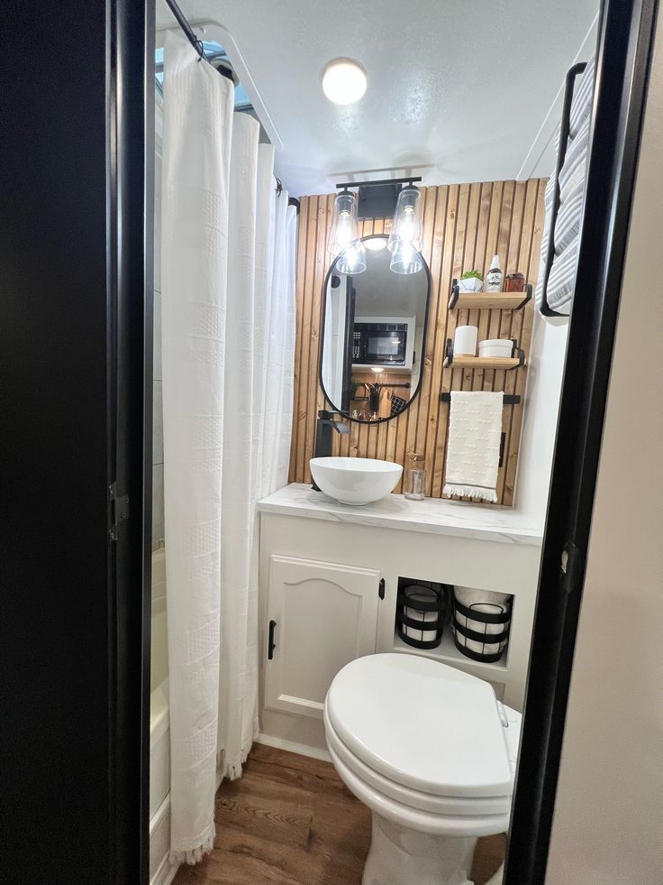 a white toilet sitting under a bathroom mirror next to a wooden floor and shower curtain