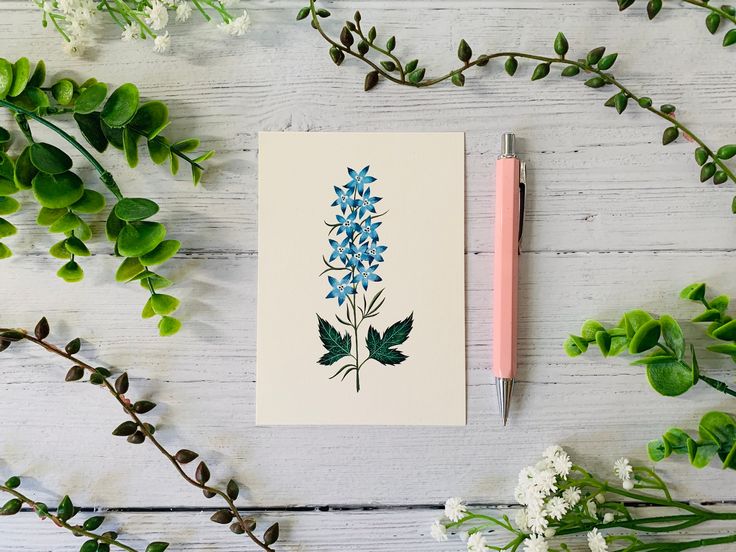 a card with blue flowers on it next to some greenery and a pen sitting on a table