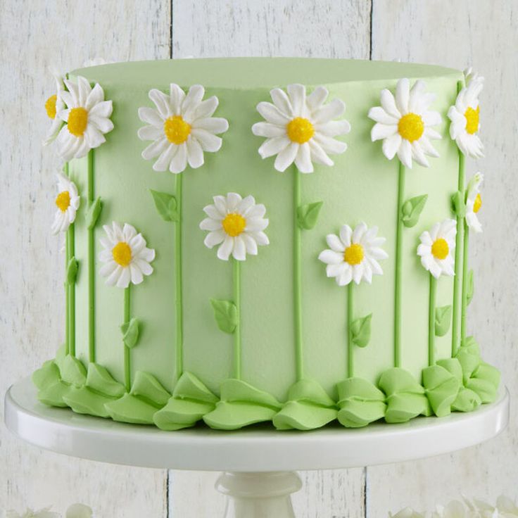 a green cake decorated with daisies and leaves on a white pedestal next to flowers