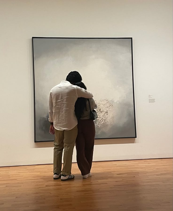 two people standing in front of a painting on the wall and looking at it with their arms around each other