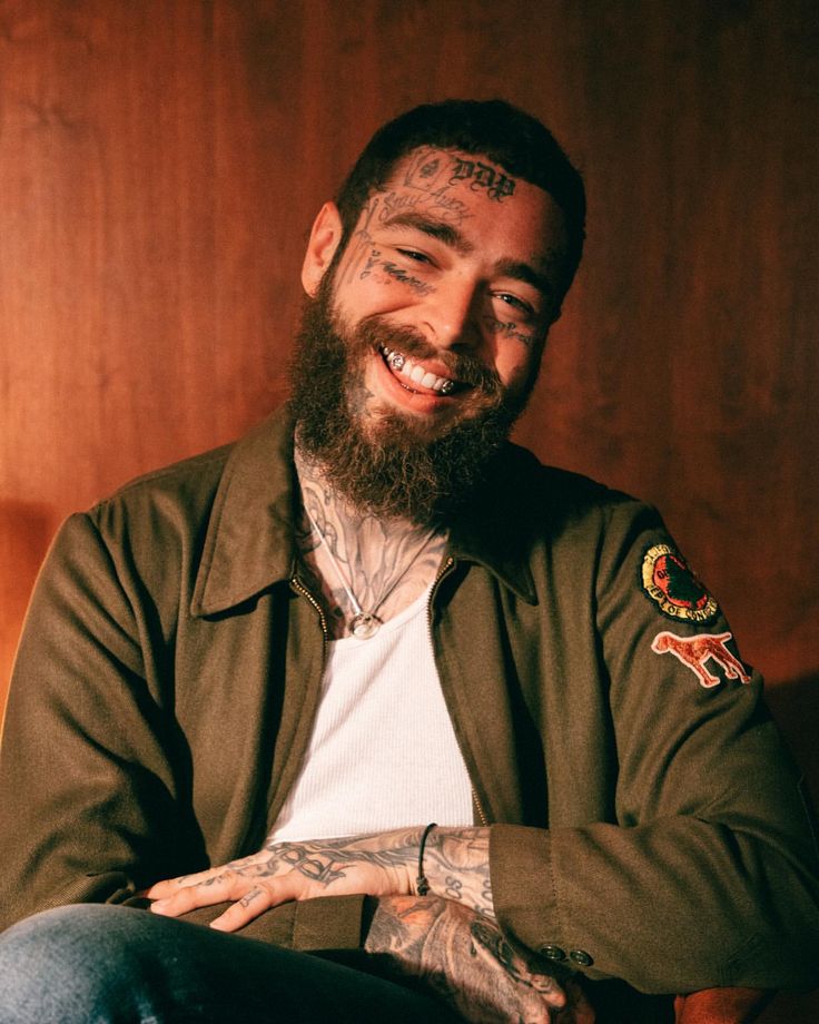 a bearded man with tattoos on his arms and chest sitting in front of a wooden wall