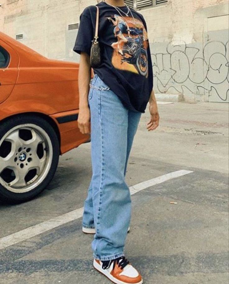 a person standing in front of a car with an orange car behind them and a building on the other side