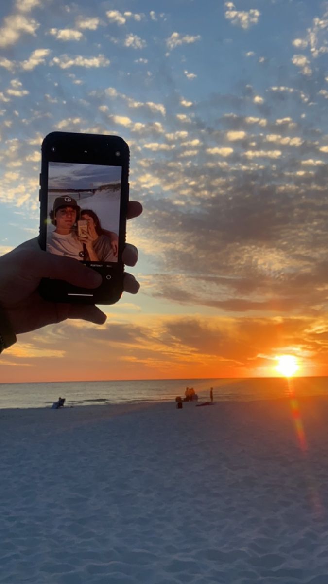 a person holding up a cell phone to take a photo on the beach at sunset