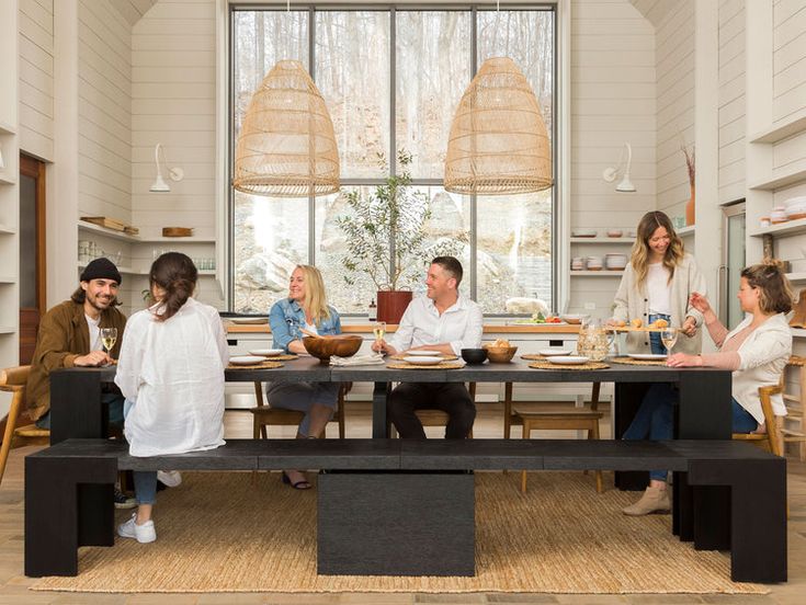 a group of people sitting around a wooden table eating food in a room with large windows